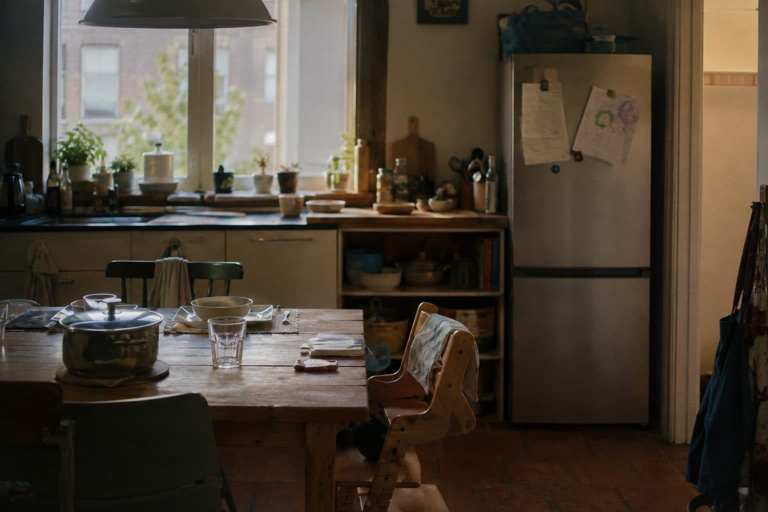 A family kitchen at the end of the day: a pan still on the table, a child's high chair pulled up to it, drawings and a letter held on the fridge door.